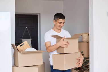 Beyaz tişörtlü, elinde karton kutuyla bekleyen, içinde eşyalarıyla birlikte paketleri boşaltan yakışıklı bir adamın kapalı mekândaki fotoğrafı..