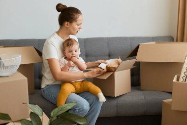 Beyaz tişörtlü üzgün bir kadının, etrafı cam kaplarla çevrili karton kutularla çevrili bir şekilde öksürük içinde otururken, yeni bir eve taşınırken, negatif duygularını ifade ederken, kucağında küçük bir kız çocuğu taşırken..