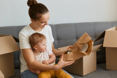 Koyu saçlı, beyaz tişörtlü beyaz bir kadının portresi kanepeye oturmuş, etrafı karton paketlerle çevrili, yeni bir daireye taşınıyor, plakalı paketleri açıyor..