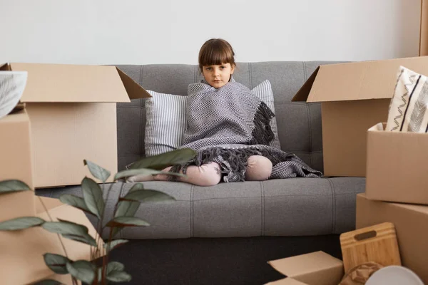 照片中可爱的心烦意乱的小女孩裹着灰蒙蒙的毯子坐在沙发上 看着相机 表达悲伤和悲伤 在重新安置的过程中感到疲惫 — 图库照片