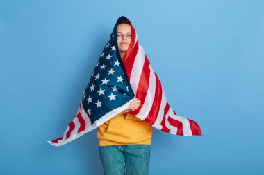 Yanağında Ukrayna bayrağı olan gururlu bir kadının portresi, Amerikan bayrağına sarılmış, kameraya bakıyor, gururunu ve desteğini hissediyor, mavi arka planda soyutlanmış bir şekilde poz veriyor..