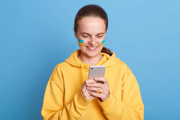 Smiling delighted woman in yellow hoodie with Ukrainian flag on cheeks, using cell phone, reading news good about war, scrolling online, posing isolated over blue background.