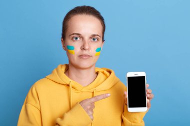Yanaklarında Ukrayna bayrağı olan kapüşonlu ciddi bir beyaz kadın, mavi arka planda izole edilmiş poz veriyor, akıllı telefona bakıyor, reklam için fotokopi alanı gösteriyor..