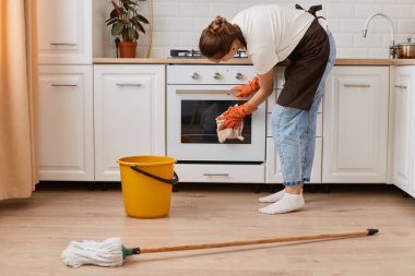 Genç yetişkin kadın portresi mutfakta paçavrayla fırını temizliyor, kadın kot pantolon, beyaz gömlek ve kahverengi önlük giyiyor, çılgın ev işleri yapıyor..