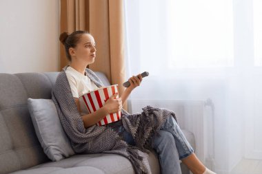 Side view portrait of beautiful young adult woman sitting on sofa in living room, watching a soap opera with sad upset facial expression, feeling sorrow.