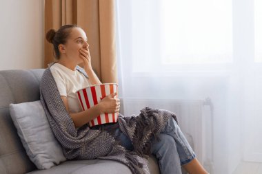 Side view portrait of young adult woman spending time at home sitting on sofa in living room, watching television, scary or horror movie, in panic eating popcorn bowl.