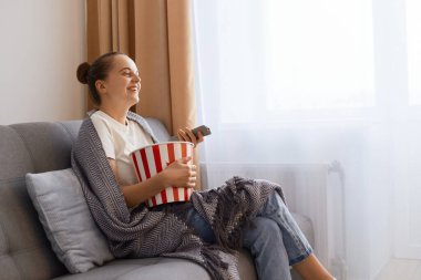 Horizontal shot of smiling happy positive woman sitting on sofa wrapped in blanket and watching tv, funny comedy show, expressing happiness, enjoying time spending at home alone.