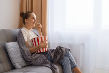 Side view photo of charming woman sitting on sofa and eating popcorn bowls, watching movie or interesting tv show, enjoying tasty snack while watch interesting tv show.