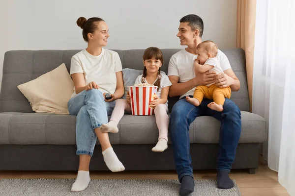 Indoor shot of satisfied family spending time together while watching tv or interesting movie, wife and husband looking at each other with smile, parents spending time with kids.