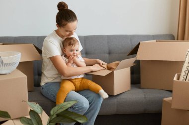Beyaz t-shirt giyen genç bir annenin kucağında küçük kızıyla kanepede otururken, eşyalarıyla birlikte kutuları açarken, yeni bir daireye taşınırken çekilmiş..