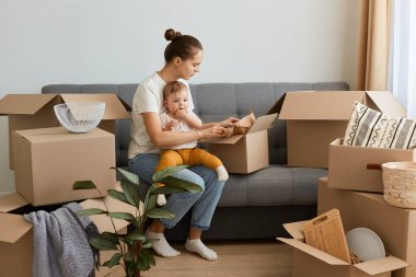 Beyaz tişörtlü, gülümseyen, siyah saçlı bir kadının portresi. Kanepede karton paketlerle çevrili ve bebeğini kucağında tutuyor..