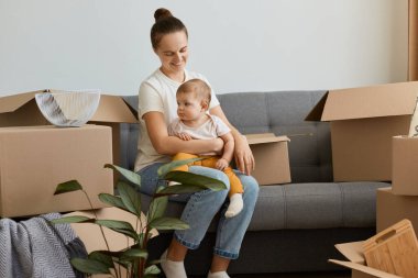 Beyaz t-shirt giyen, kanepenin üzerinde oturan, bebeğini kucağında tutan, yeni bir daireye taşınan, mutluluğunu ifade eden mutlu bir kadın..