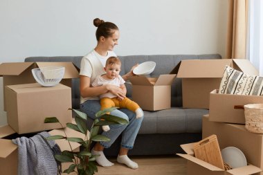 Pozitif siyah saçlı, beyaz tişörtlü bir kadının uzun bir portresi. Kanepenin üzerinde karton kutularla çevrili bir şekilde oturuyor ve kızını elinde tutuyor. Tabaklarla kutuları açıyor..