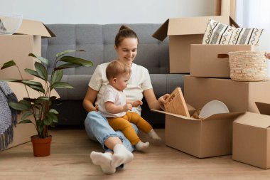 Koyu renk saçlı, gündelik giysiler giyen, kızıyla birlikte yerde oturan ve karton kutulardan kişisel eşyaları boşaltan mutlu bir kadın..