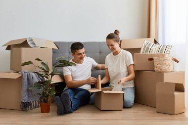 Happy Caucasian family moved in new apartment, sitting on the floor among cardboard boxes, couple in love rent a house together, relocated in new home, unpacking personal belongings.