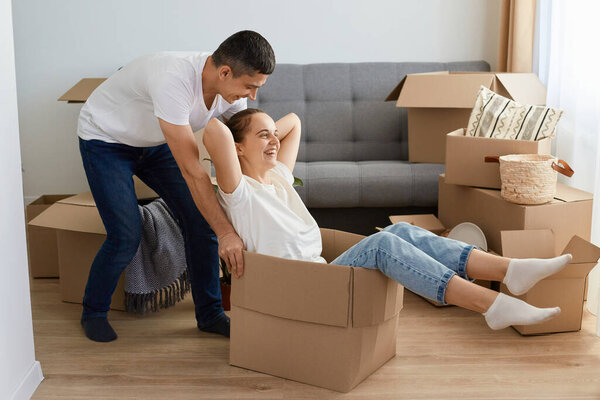 Portrait of positive delighted couple having fun laughing, relocating into new apartment, woman riding in cardboard box while man pushing it, family unpacking personal piles together.