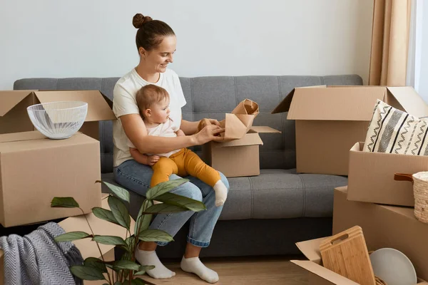 Yatay olarak gülümseyen, tatmin olmuş beyaz tişörtlü bir kadının kanepeye oturup kutuları açtığı, yeni bir eve taşındığı ve küçük kızıyla birlikte poz verdiği bir fotoğraf..