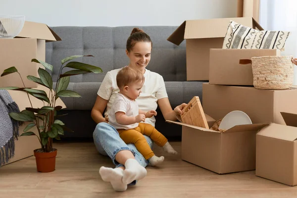 Koyu renk saçlı, gündelik giysiler giyen, kızıyla birlikte yerde oturan ve karton kutulardan kişisel eşyaları boşaltan mutlu bir kadın..