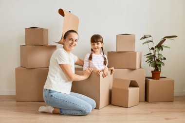 Beyaz tişört ve kot pantolon giyen, kızıyla birlikte yerde oturan, kameraya bakan, yeni bir eve taşınırken karton paketlerle oynayan bir kadın..