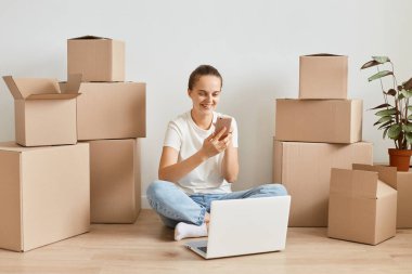 Kot pantolon ve beyaz t-shirt giyen mutlu, yetişkin bir kadının karton kutuların yanında oturduğu, telefon ettiği, dizüstü bilgisayarda çalıştığı ve olumlu duygular ifade ettiği kapalı çekimler..