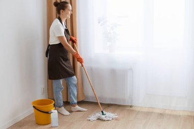 Genç bir kadının oturma odasında yerleri paspas ile yıkarken, ayakta durup pencereye bakarken, beyaz tişört, kahverengi önlük giyerken, evi temizlerken olumlu duygularını ifade ederken çekilmiş bir fotoğrafı..
