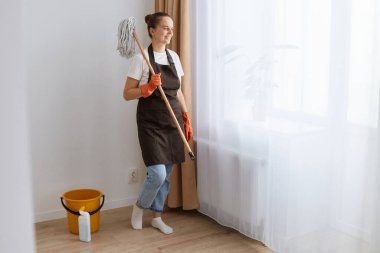 Çekici, genç, yetişkin bir ev kadınının evini temizlemesi, enfeksiyon güvenliği, paspas tutması, temizlik, eldiven, önlük, tişört ve kot giymesi, pencerenin yanında durması..