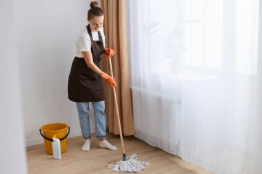Genç yetişkin bir kadının portresi oturma odasının yanında paspasla yerleri yıkıyor, paspasla el ele tutuşuyor, modern bir dairede temizlik hizmeti veriyor..