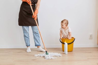 Kimliği belirsiz bir kadının önlük giydiği, kot pantolon giydiği, paspas ile yerleri sildiği, bebek kızının kovada oturduğu, anneyle vakit geçirdiği,.