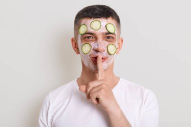 Beyaz tişört giyen yakışıklı bir adamın portresi kameraya bakıyor, yüzünde maske ve salatalık var, parmağını ağzının yakınında tutuyor, sırrını gri arka planda saklı tutmak istiyor..