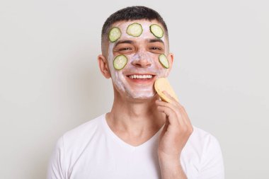 Beyaz tişört giyen yakışıklı bir adam maske, salatalık ve sünger dilimleri kullanarak cilt bakımı yapıyor. Gri arka planda izole edilmiş bir şekilde kameraya bakıyor..