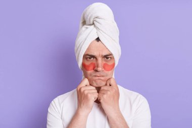 Yapışkan hidrojel göz bantlı üzgün kızgın adamın kapalı çekim, kozmetik işlemler yapıyor, göz altı kolajen bantlı kameraya bakıyor ve başının üstünde havlu var..