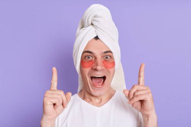 Beyaz t-shirt giyen heyecanlı bir adamın portresi ve kafaya sarılmış havlu, göz altı yamalarla poz vermek, yukarı işaret etmek, cilt bakımı prosedürleri hakkında yeni bir fikir, mor arka planda izole edilmiş.
