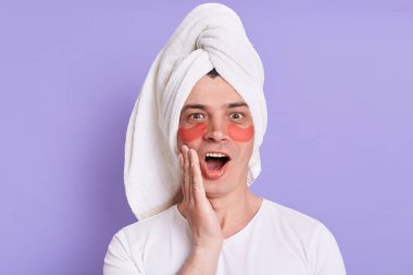 Şaşkın adam tişört giyer ve kafaya havlu sarar, göz altı yamalarıyla poz verir, ağzını kapalı tutar, mor arka planda izole edilmiş yeni kozmetik ürünü anons eder..