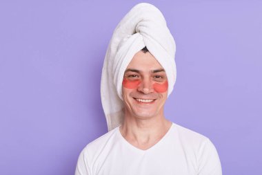 Beyaz t-shirt giyen ve kafaya havlu sarılmış memnun bir adam, olumlu duygularını ifade eden, sabah kozmetoloji prosedürleri yapan, mor arka planda izole edilmiş göz altı yamalarıyla poz veren.