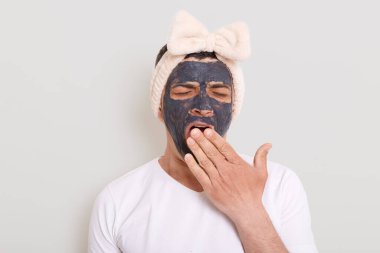 Beyaz tişört giyen komik bir adamın portresi ve kadın saç bandı çamur maskesiyle ayakta duruyor ve esniyor, palmiye ile ağzını örtüyor, beklemekten yoruluyor ve gri arka planda izole edilmiş bir şekilde duruyor..