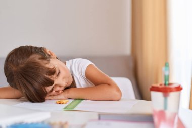 Beyaz tişört giyen, masa başında uyuyan, ödevlerini yaparken yorgun, masasında uzanmış, yorgun ve aşırı çalışılmış, siyah saçlı bir çocuğun kapalı çekimleri..