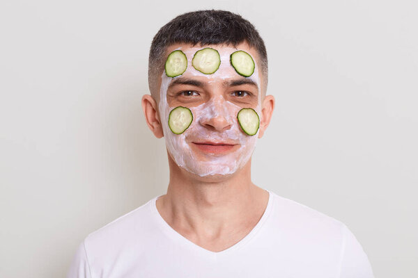 Indoor shot of smiling positive man wearing white t shirt, standing looking at camera with mask and slices of cucumbers on his face, posing isolated over gray background.