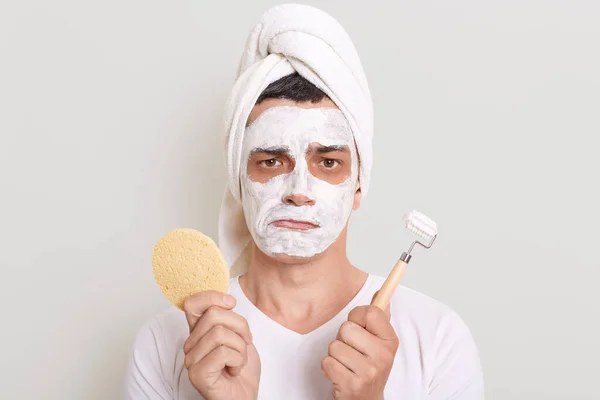 Sad man wearing white t shirt and towel on head posing isolated over gray background, holding sponge and massage roller in hands, doing skin care procedures, standing with frowning face.