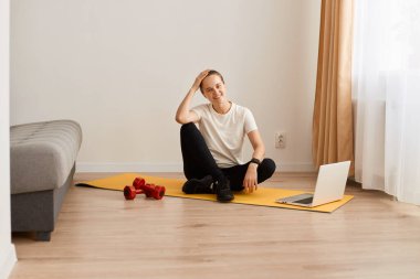 Portrait of tired young adult woman sitting on yoga mat with happy facial expressing after online training with trainer, female with sporty body wearing white t shirt and leggins.