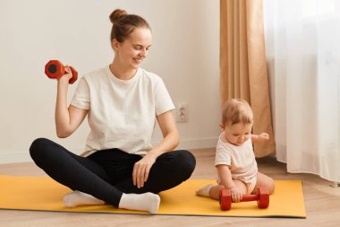 Küçük kızıyla yoga minderinde oturup, kollarını, pazılarını ve trisepslerini çalıştırırken spor egzersizleri yaparken ve yeni yürümeye başlayan çocuğuna bakarken çekilmiş gülümseyen bir fotoğraf..