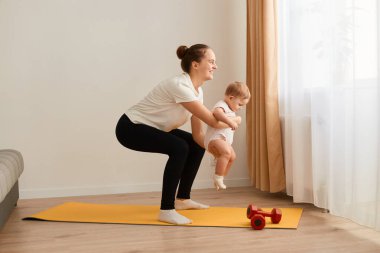 Side view portrait of young adult mother in t shirt and leggins doing sit ups with her baby in hands, looking after of kid and doing workout for her body.