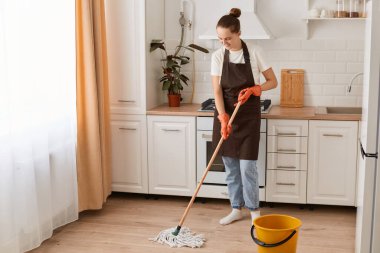 Indoor shot of happy Caucasian woman housewife cleaning floor with mop in kitchen, wearing white t shirt, jeans and brown apron, expressing positive emotions while washing her flat.