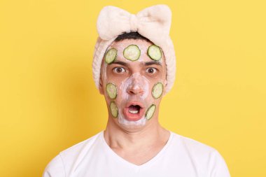 Beyaz tişörtlü ve saç bantlı şaşkın adam, yüz maskesi ve salatalık ile poz sarı arka planda izole edilmiş, şaşırmış ifade, büyük gözler ve ağız kameraya bakarak