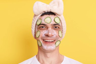 Beyaz tişört giyen, fiyonklu saç bandı takan, yüz maskesi ve salatalıkla poz veren, kameraya bakan, güzellik prosedürlerinden memnun, sarı arka planda izole edilmiş mutlu bir adam..