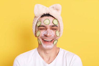 Beyaz tişört ve fiyonklu saç bandı takan, sarı arka planda saklanmış yüz maskesi ve salatalıkla poz veren mutlu pozitif bir adam. Gülmek, kozmetoloji işlemleri yapmak için çok komik..