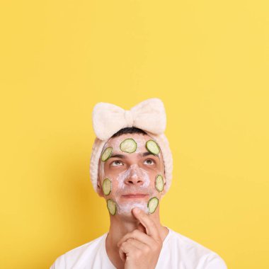 Beyaz tişört ve fiyonklu saç bandı takan, sarı arka planda gizlenmiş yüz maskesi ve salatalıkla poz veren, fotokopi alanına bakan, spa salonu reklamına bakan dalgın bir adam..