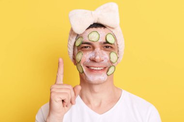 Olumlu iyimser genç adam beyaz t-shirt ve kurdeleli saç bandı takıyor, sarı arka planda izole edilmiş yüz maskesi ve salatalık takıyor, fotokopi alanını işaret ediyor..