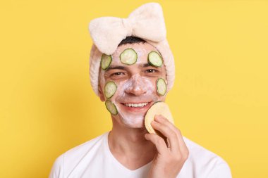 Beyaz tişört giyen, fiyonklu saç bandı takan, yüz maskesi ve salatalıkla poz veren, kozmetik işlemler yapan, sünger kullanan, sarı arka planda izole edilmiş,.