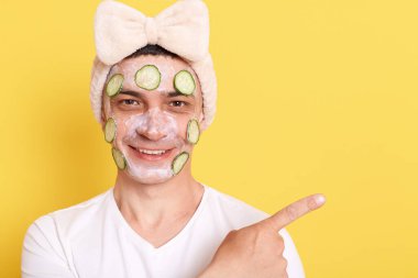 Gülümseyen, beyaz tişört giyen, fiyonklu saç bandı takan, reklam için bölgeyi işaret eden, sarı arka planda izole edilmiş yüz maskesi ve salatalıkla poz veren mutlu adam..