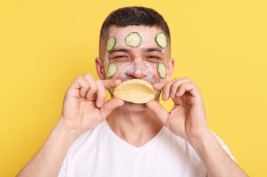 Gülümseyen komik adam maskesini salatalıkla takıyor, sünger tutuyor ve ağzını kapatıyor, kameraya olumlu bir ifadeyle bakıyor, sarı arka planda izole bir şekilde poz veriyor..
